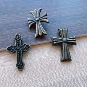 以下が含まれることがあります： さまざまなデザインとサイズの3つの装飾的な金属製の十字架。十字架はアンティークブロンズ仕上げで、木製の表面に配置されています。デザインには、装飾的なディテールと、古典的な十字架の形が含まれています。