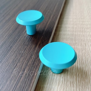 May include: Two turquoise cabinet knobs with a rounded top and a cylindrical base. The knobs are set against a dark brown and light brown wood grain surface. The knobs are a bright, solid colour.