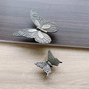 May include: Two decorative metal butterfly drawer pulls in a silver finish. The larger butterfly has a more intricate design with a detailed wing pattern. The smaller butterfly has a simpler design with a more rounded wing shape.
