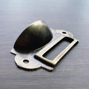 Puede incluir: Tirador de cajón de latón antiguo con parte superior redondeada y un portaetiquetas rectangular. El portaetiquetas tiene un borde elevado.