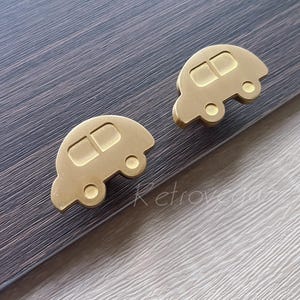 Op de afbeelding: Twee goudkleurige, auto-vormige kastknoppen. Elke knop heeft twee rechthoekige ramen en twee cirkelvormige wielen. De knoppen zijn van metaal en staan tegen een donkere houtnerf achtergrond.