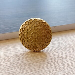 Puede incluir: Un pomo de metal dorado con un diseño circular que presenta un patrón repetitivo de cuadrados y círculos. El pomo tiene un acabado liso y pulido.