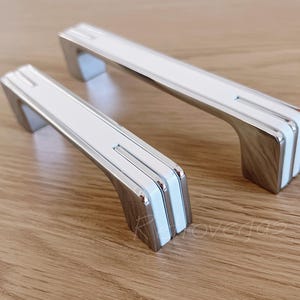 Puede incluir: Dos tiradores de armario blancos y cromados con un diseño moderno. Los tiradores tienen una forma rectangular con bordes redondeados y un diseño de rayas blancas.