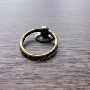 May include: A gold-toned ring pull cabinet handle with a round base. The handle is on a brown wooden surface.