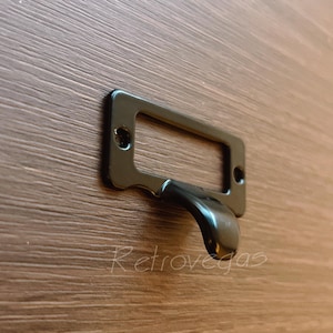 Puede incluir: Porta etiquetas de metal negro con una abertura rectangular y un fondo curvo. El porta etiquetas está fijado a una superficie de madera.