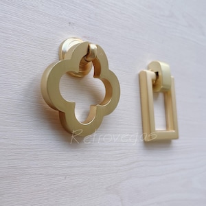 May include: Two gold-toned cabinet pulls. One is a four-leaf clover shape with a cutout in the center. The other is a rectangular pull with a cutout in the center.