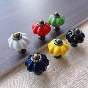 May include: Six ceramic pumpkin-shaped drawer knobs in various colors, including red, green, white, yellow, blue, and black. Each knob has a gold-colored metal accent.