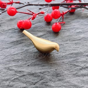 May include: A gold-colored metal bird figurine on a gray textured surface. The bird is facing right and has a slightly curved beak.