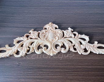 May include: A carved wooden decorative architectural element with an ornate design. The light-colored wood features a central oval shape flanked by symmetrical floral and scrollwork patterns. The piece is designed to be mounted on a surface.