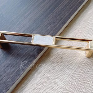 May include: Gold cabinet door pull with a rectangular white inlay. The pull has a rectangular frame with a rectangular opening in the centre.