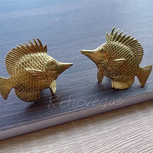 May include: Two gold-coloured metal fish figurines with detailed scales and fins. The fish are facing opposite directions and have a slightly raised, textured surface.