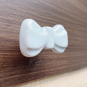 Może przedstawiać: Biały ceramiczny uchwyt do szuflady w kształcie muszki. Muszka ma prosty design bez ozdób.