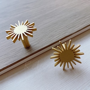 以下が含まれることがあります： ゴールドの太陽の形をしたキャビネットノブが2つ。ノブは、隆起した円形のセンターと放射状のポイントを持っています。