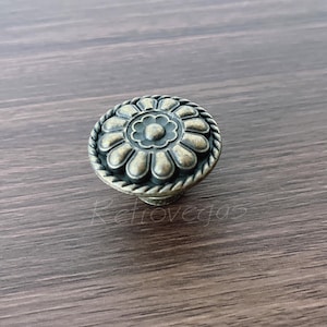 May include: Antique brass cabinet knob with a floral design. The knob features a central flower surrounded by petal-shaped details and a rope-like border. The hardware is made of metal and has a vintage aesthetic.