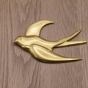 May include: A gold-colored metal sculpture of a swallow in flight. The sculpture is on a woodgrain background.