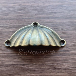 May include: A vintage-style, brass-coloured cabinet pull with a scalloped design. The pull is shaped like a half-circle and has a smooth, slightly textured surface.