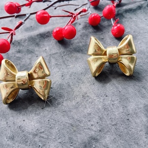 Könnte beinhalten: Zwei goldfarbene Ohrringe in Form einer Schleife. Die Schleifen sind aus Metall und haben eine glänzende Oberfläche.