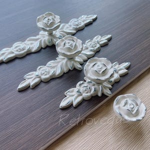 May include: White rose-shaped cabinet hardware with silver trim. The set includes three pulls and one knob. The floral design is set against a dark gray wood-grain surface, creating a vintage aesthetic. Ideal for furniture restoration or adding a touch of elegance.
