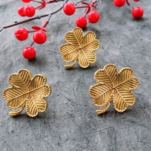 May include: Three gold-colored four-leaf clover charms. The charms are detailed with a textured design and have a stem.