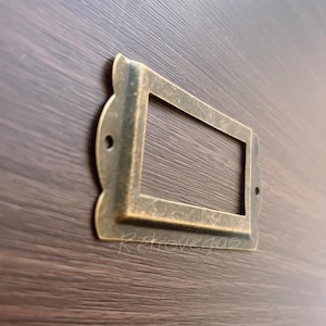 Puede incluir: Un portatarjetas de latón de estilo vintage con una abertura rectangular y bordes decorativos. El soporte está montado sobre una superficie de vetas de madera oscura. La palabra "Retrovegas" es visible.