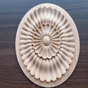 K&ouml;nnte beinhalten: Oval geformte, unfertige Holzschnitzerei mit detailliertem Blumendesign. Das zentrale Element ist ein kreisf&ouml;rmiges Muster mit bl&uuml;tenblattartigen Formen, die sich nach au&szlig;en erstrecken. Der &auml;u&szlig;ere Rand des Ovals hat ein Muscheldesign. Das Holz hat eine nat&uuml;rliche, helle Farbe.