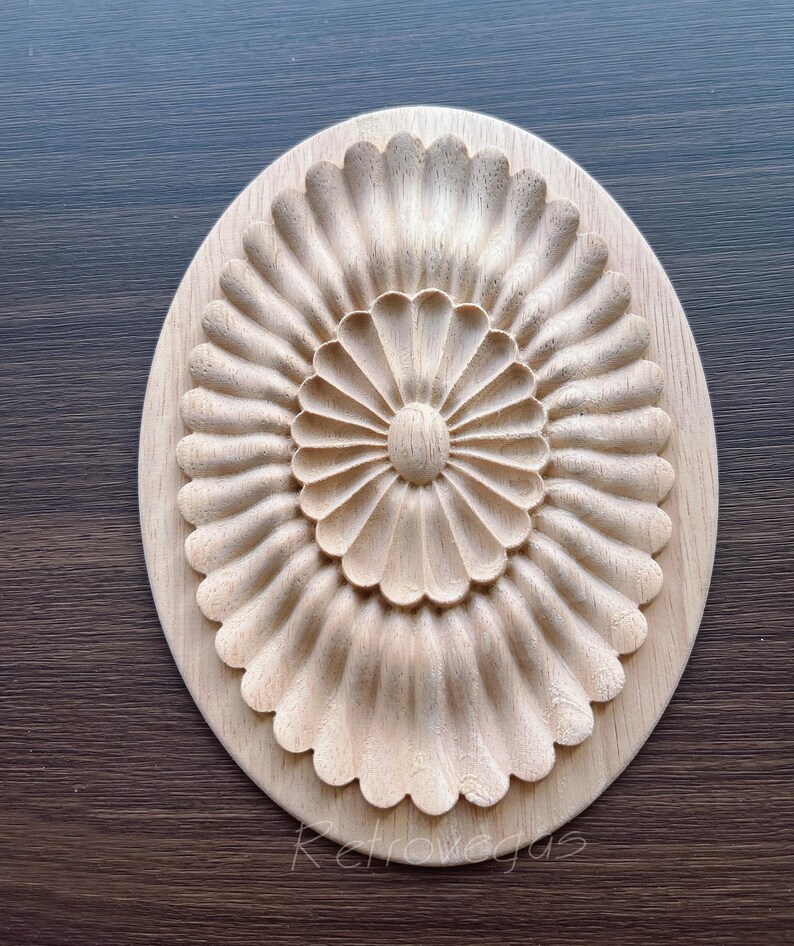 K&ouml;nnte beinhalten: Oval geformte, unfertige Holzschnitzerei mit detailliertem Blumendesign. Das zentrale Element ist ein kreisf&ouml;rmiges Muster mit bl&uuml;tenblattartigen Formen, die sich nach au&szlig;en erstrecken. Der &auml;u&szlig;ere Rand des Ovals hat ein Muscheldesign. Das Holz hat eine nat&uuml;rliche, helle Farbe.