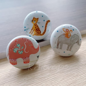 Peut inclure: Trois boutons en céramique avec des illustrations d'animaux. Le premier bouton représente un éléphant rose avec un motif vert et blanc. Le deuxième bouton représente un éléphant gris avec un singe monté sur son dos. Le troisième bouton représente un léopard jaune avec des taches noires.