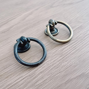 Puede incluir: Dos tiradores de anillo de estilo antiguo, uno en negro y otro en dorado. Ambos tienen un anillo redondo y una perilla decorativa.