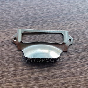 Peut inclure: Une poignée de tiroir en métal argenté avec un porte-étiquette rectangulaire. La poignée a une forme incurvée et est conçue pour être utilisée sur des armoires ou des tiroirs.