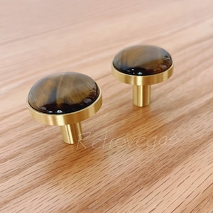 Op de afbeelding: Twee kastknoppen met een ronde tijgeroogstenen bovenkant en een goudkleurige basis. De knoppen staan op een licht houten oppervlak. De knoppen hebben een klassiek, elegant ontwerp.