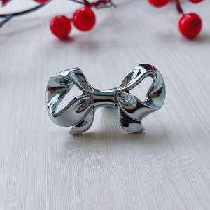 Puede incluir: Un pomo de armario o tirador de cajón en forma de lazo plateado. El lazo está hecho de metal y tiene un acabado brillante.