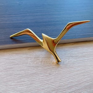 May include: A gold-colored metal bird sculpture with a stylized design. The bird is in flight with its wings spread out.