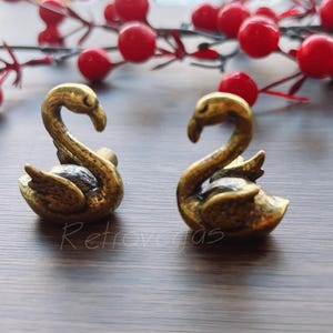 Op de afbeelding: Twee kleine, antiek goudkleurige zwaanbeeldjes. De zwanen staan tegenover elkaar, met hun halzen gebogen. Rode bessen en takken zijn op de achtergrond. Het woord "Retrovegas" is cursief op het oppervlak geschreven.