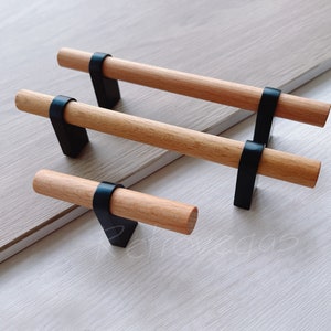 May include: Three wooden cabinet pulls with black metal hardware. The pulls are different sizes and are arranged in a row on a light wood surface.