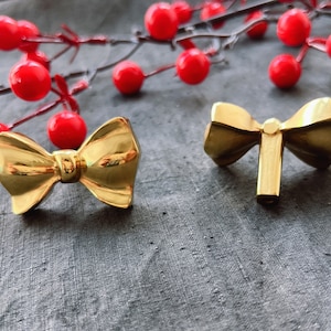 May include: Two gold bow-shaped door knobs on a grey fabric background. The knobs are made of metal and have a shiny finish.