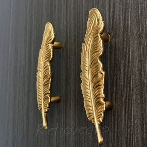 Pode incluir: Dois puxadores de armário em forma de pena dourados. Os puxadores são detalhados com um design de pena texturizado.