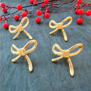 May include: Four gold-coloured bow-shaped drawer pulls. The pulls are arranged in a square pattern on a grey surface.