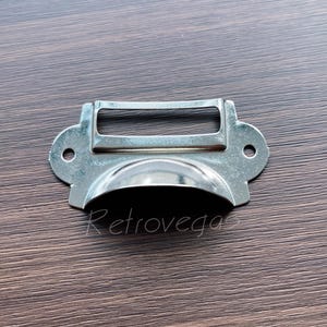 May include: Silver metal drawer pull with a rectangular label holder. The pull has a rounded, slightly curved shape.