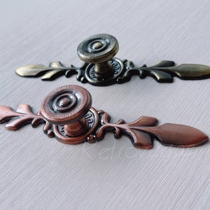 May include: Two ornate cabinet knobs with a decorative floral design. The knob on the left is copper colored and the knob on the right is gold colored.