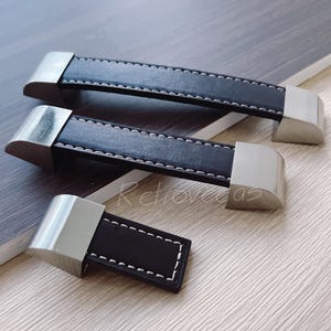 May include: Three black leather cabinet pulls with silver metal accents. The pulls have a stitched edge and are different sizes.