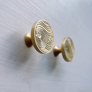 Op de afbeelding: Twee gouden kastknoppen met een ronde basis en een reliëfdesign van draaiende lijnen. De knoppen zijn gemonteerd op een witte muur.