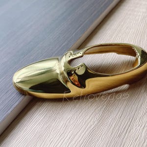 Könnte beinhalten: Ein goldfarbener, schuhförmiger Schlüsselhalter aus Metall. Der Schlüsselhalter ist hohl und hat eine glatte, glänzende Oberfläche.