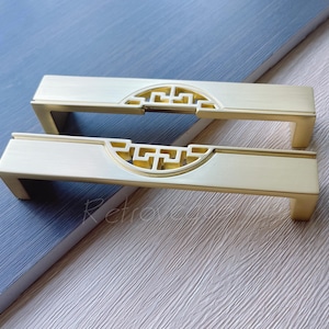 May include: Two brass-toned cabinet pulls with a rectangular design and a decorative cutout pattern. The pulls have a modern aesthetic and are set against a textured background.