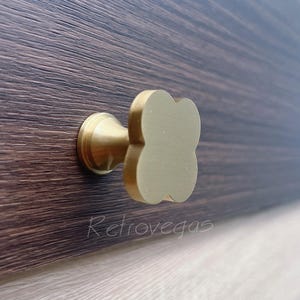 May include: A gold-toned cabinet knob with a four-leaf clover design. The knob is mounted on a dark wood surface.