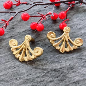 May include: Two gold-toned decorative cabinet pulls with a fan-shaped design. The pulls have a detailed, intricate design.