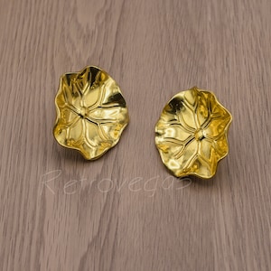 Könnte beinhalten: Ein Paar goldfarbene Ohrringe mit einem blütenförmigen Design. Die Ohrringe sind aus Metall und haben eine strukturierte Oberfläche.