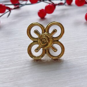 May include: Gold-toned metal cabinet knob with a decorative rose design. The knob has a four-leaf clover shape with a circular center.