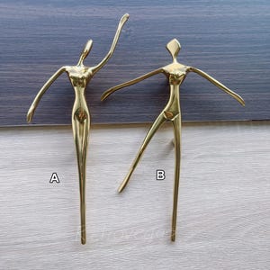 Peut inclure: Deux sculptures en métal doré représentant des danseurs stylisés. L'un des danseurs a les bras levés, l'autre a une jambe tendue. Les sculptures sont étiquetées A et B. Le fond est un grain de bois foncé.