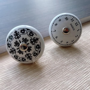 以下が含まれることがあります： 黒いデザインが施された白いセラミック製のノブが2つ。1つのノブには、黒いローマ数字と「パリ」の文字が書かれた時計の文字盤があります。もう1つのノブには、黒い花柄のデザインが施されています。