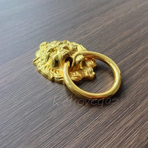 Puede incluir: Un llamador de puerta de cabeza de león dorado con un anillo redondo. La cara del león está detallada con una melena y una boca cerrada.